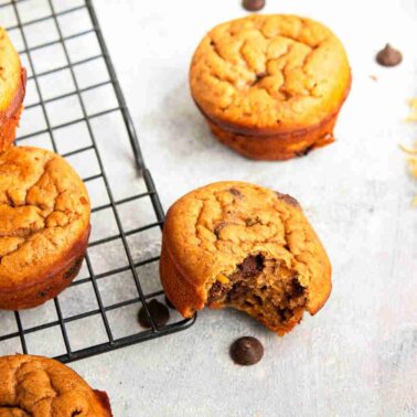 Several chocolate chip muffins are on a cooling rack, with one bitten muffin off to the side. Perfect as healthy snacks to keep hands busy, a few chocolate chips and a yellow napkin complete the tempting scene.