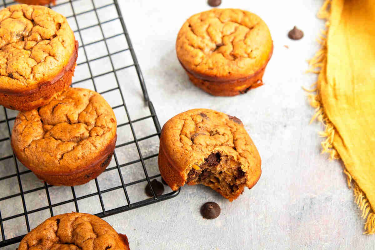 Several chocolate chip muffins are on a cooling rack, with one bitten muffin off to the side. Perfect as healthy snacks to keep hands busy, a few chocolate chips and a yellow napkin complete the tempting scene.
