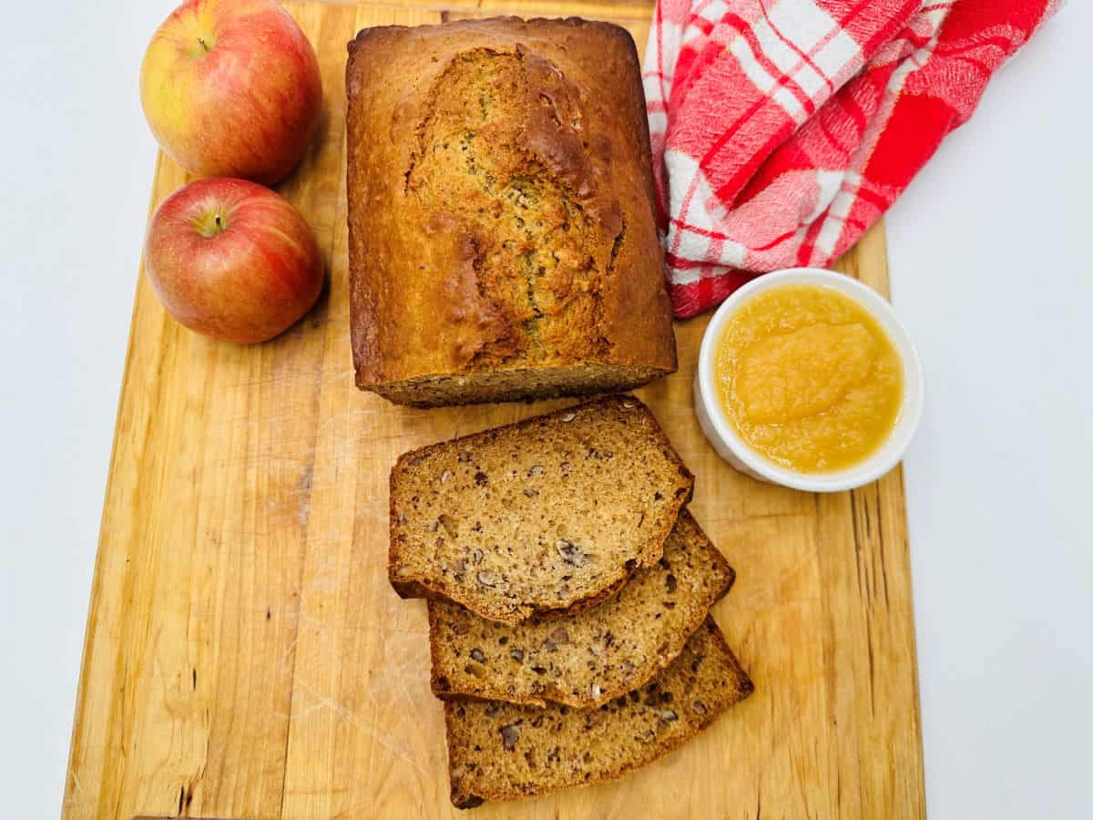 A loaf of banana bread with three slices cut, two apples, a bowl of applesauce, and a red checked towel on a wooden board—perfect for fall recipes that celebrate cozy nights and crisp air.