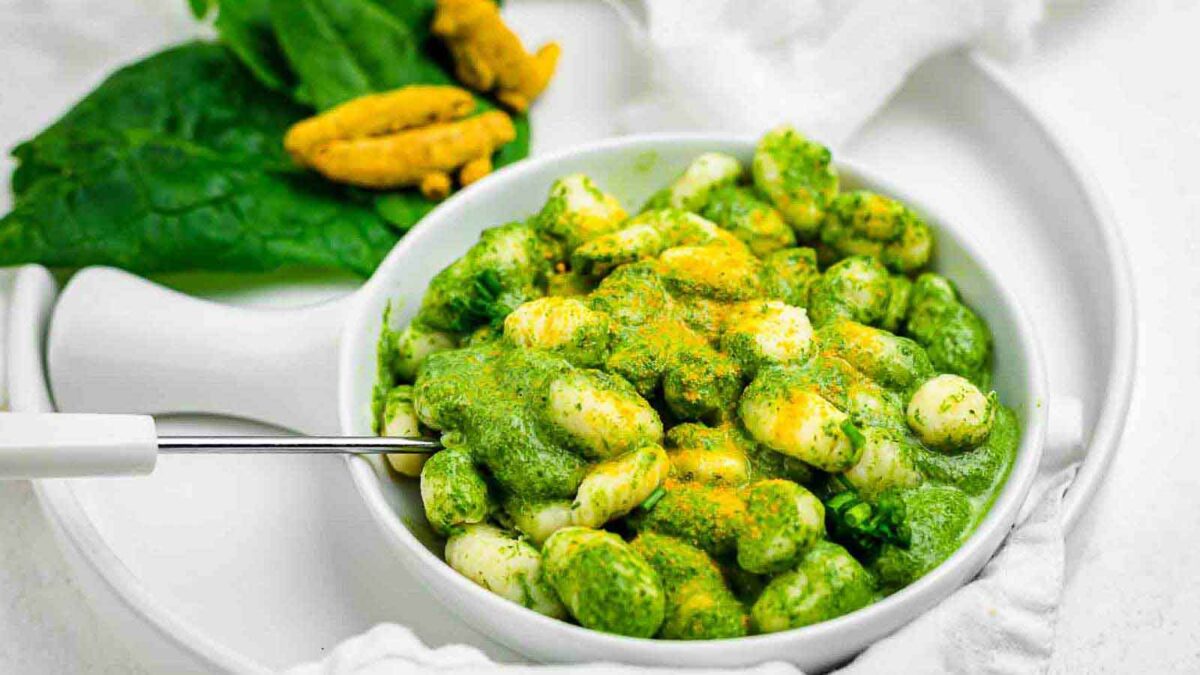 A white bowl of gnocchi topped with green sauce and yellow seasoning, placed on a white plate with green leaves and yellow flowers—perfect for cozy nights when fall recipes warm you up against the crisp air.