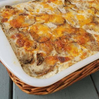 A white casserole dish filled with baked scalloped potatoes, topped with melted cheese, sits on a wooden surface—perfect for cozy nights and enjoying the crisp air of fall.