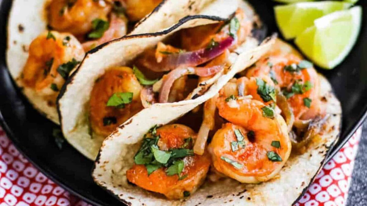 Three shrimp tacos topped with cilantro and onions on corn tortillas, served with lime wedges on a black plate—perfect for easy dinners and busy weeknights or anyone craving quick seafood recipes.
