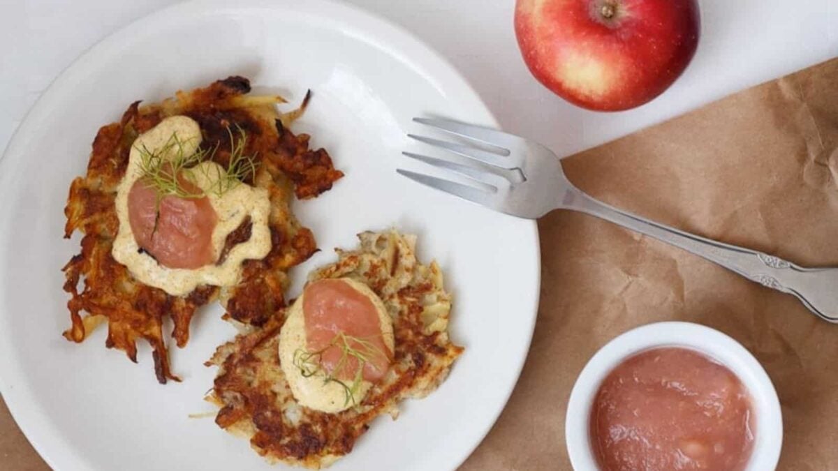 Two potato pancakes topped with sauce and garnish on a white plate, perfect for Kids Breakfasts. A fork, an apple, and a small bowl of pink sauce add to this tasty Back-To-School Breakfasts idea.