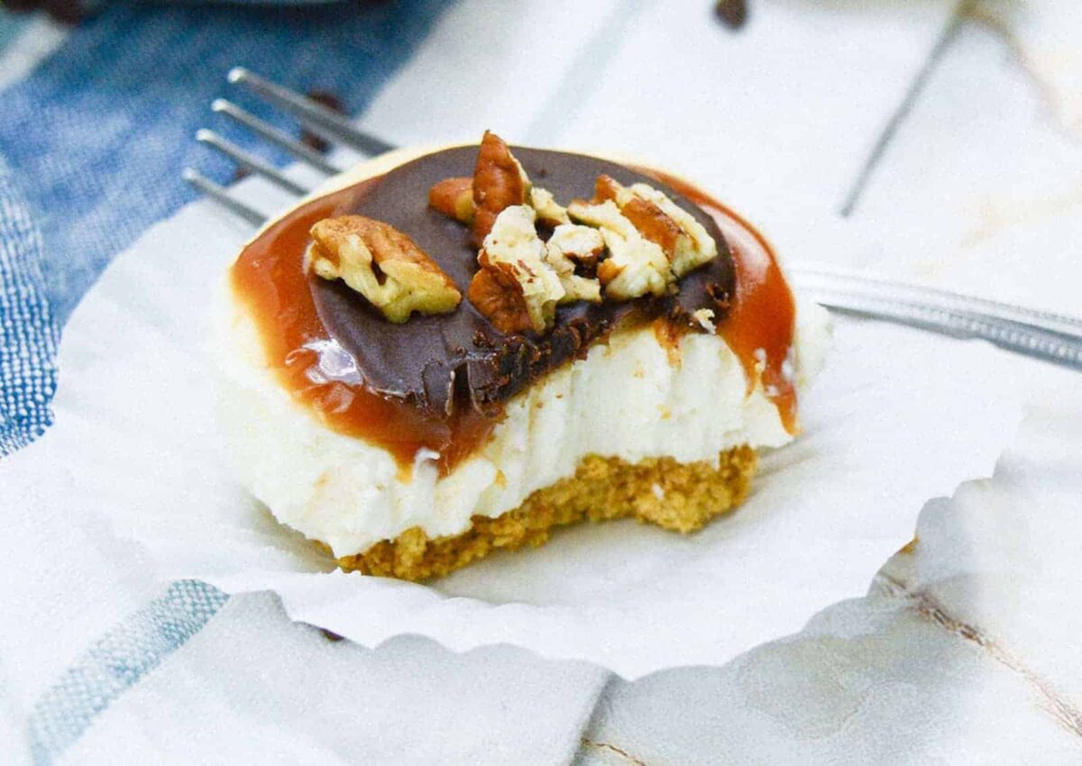 A partially eaten cheesecake square with a graham cracker crust, chocolate and caramel topping, and pecan pieces sits on a white paper liner with a fork nearby—perfect for Desserts at potlucks, as these treats travel well.