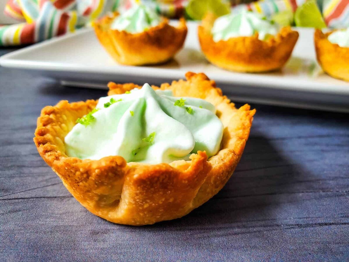 Mini tart with a golden, flaky crust filled with whipped cream and green zest, placed on a dark surface with more tarts in the background—these make perfect desserts for potlucks or any occasion needing desserts that travel well.