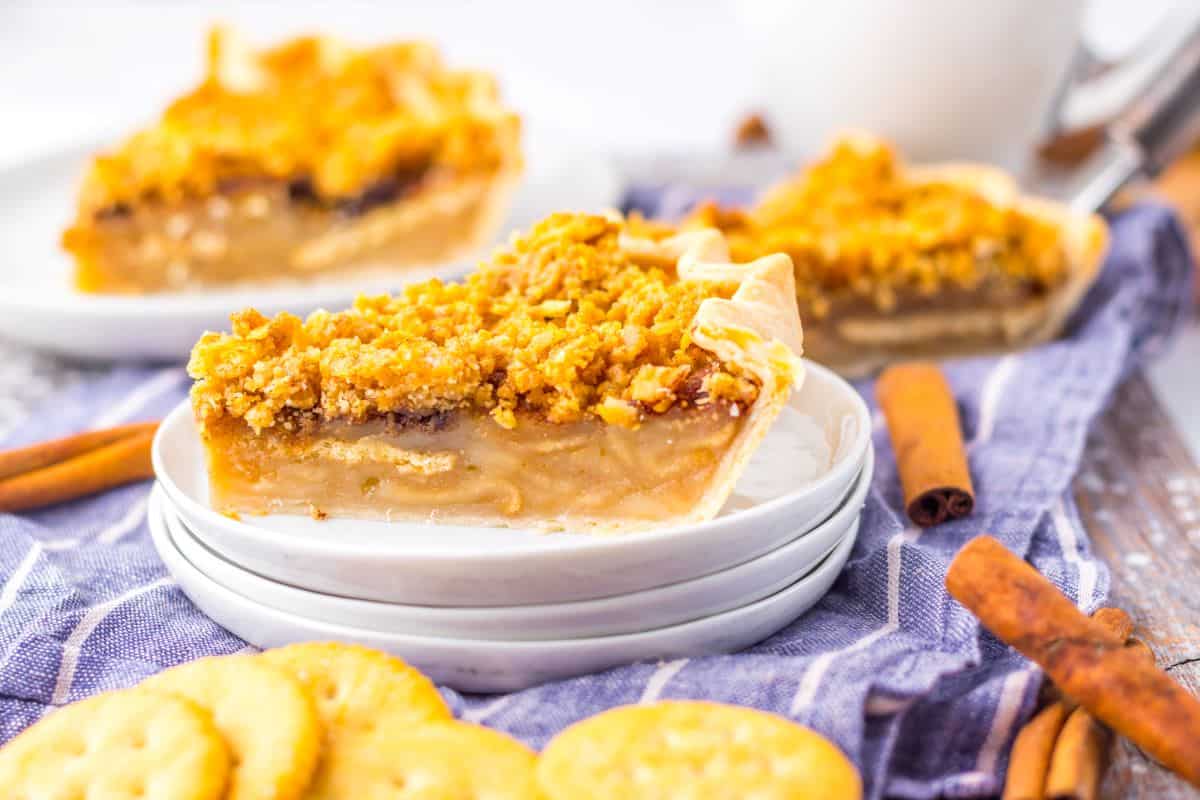 A slice of pie with crumb topping sits atop a stack of white plates—perfect potluck desserts—surrounded by crackers, cinnamon sticks, and more pie in the background.