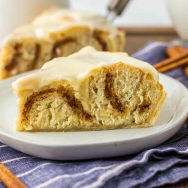 A slice of cinnamon roll with icing on a white plate, placed on a blue striped cloth with cinnamon sticks nearby—one of the best desserts that travel well and perfect for potluck desserts.