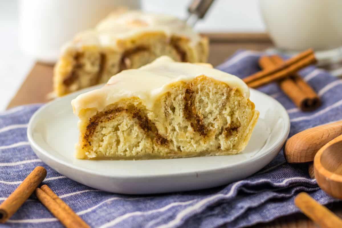 A slice of cinnamon roll with icing on a white plate, placed on a blue striped cloth with cinnamon sticks nearby—one of the best desserts that travel well and perfect for potluck desserts.