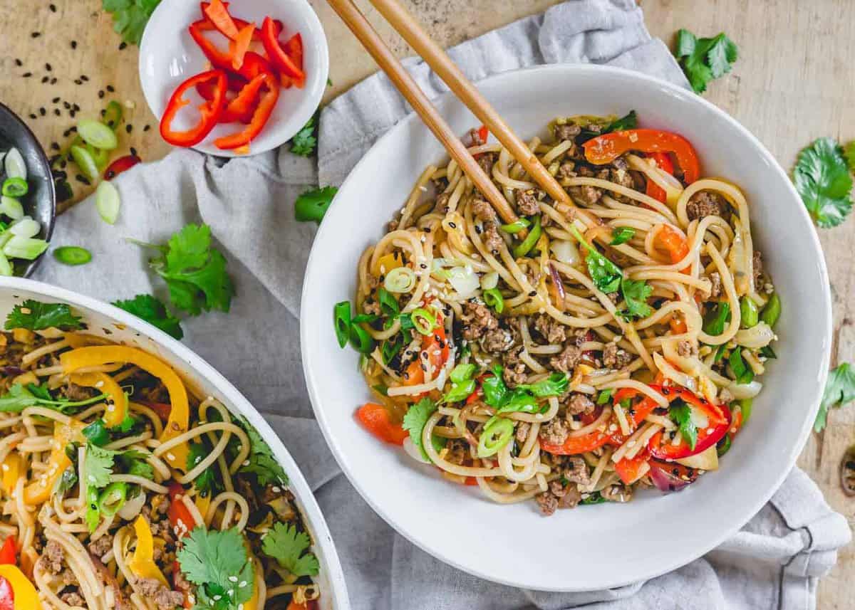 A bowl of easy stir-fry noodles with ground meat, colorful bell peppers, green onions, and cilantro, chopsticks resting on the bowl; perfect for weeknight recipes with extra toppings on the side.