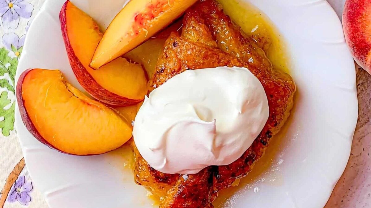A slice of peach cobbler on a white plate, topped with whipped cream and served with fresh peach slices—a classic among desserts made with few ingredients.