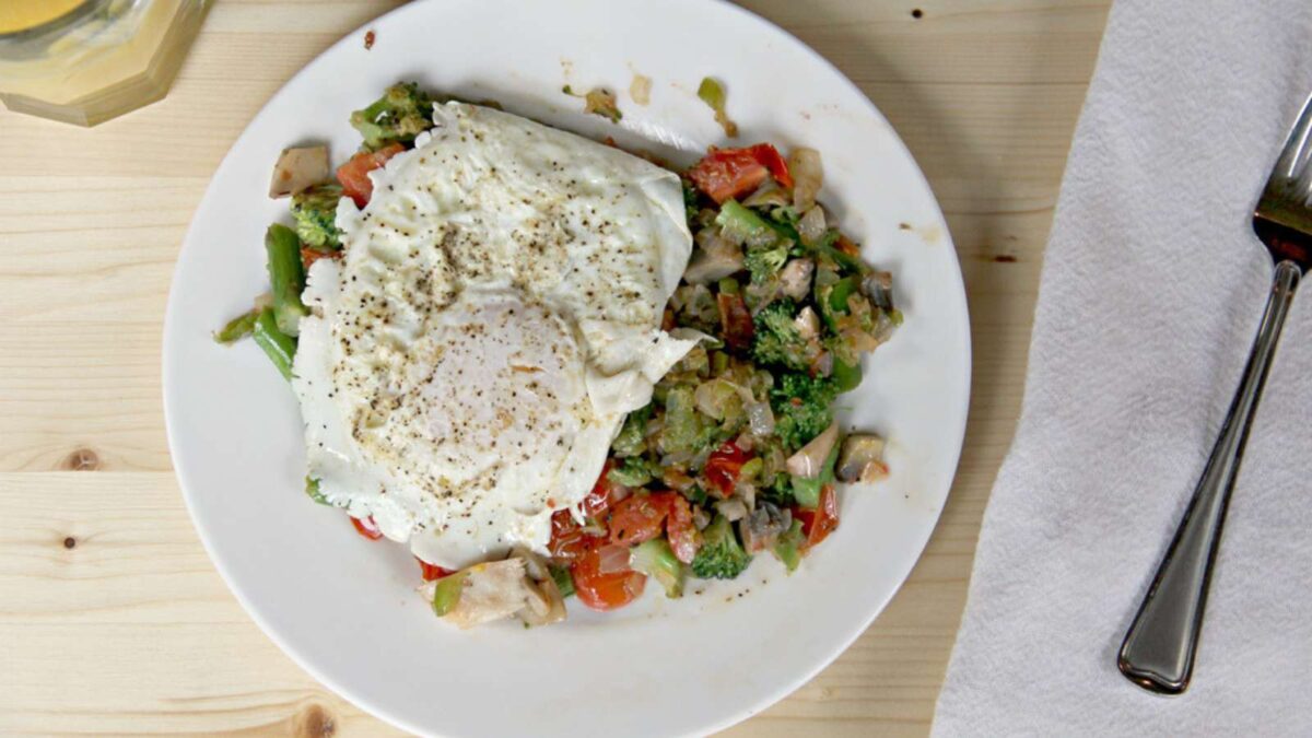 A plate of stir-fried vegetables topped with a fried egg, next to a fork and a folded napkin on a light wooden table—perfect inspiration for egg breakfast recipes.