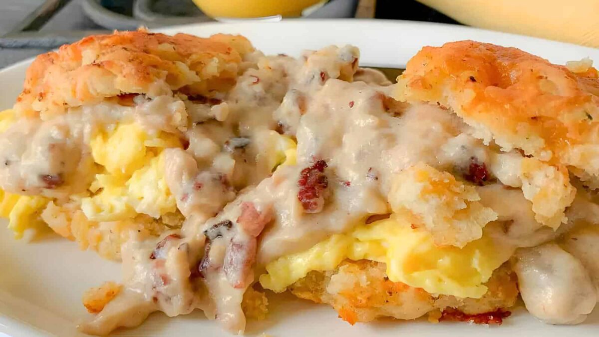 A biscuit sandwich filled with scrambled eggs and topped with sausage gravy on a white plate—perfect for those seeking delicious Egg Breakfast Recipes.