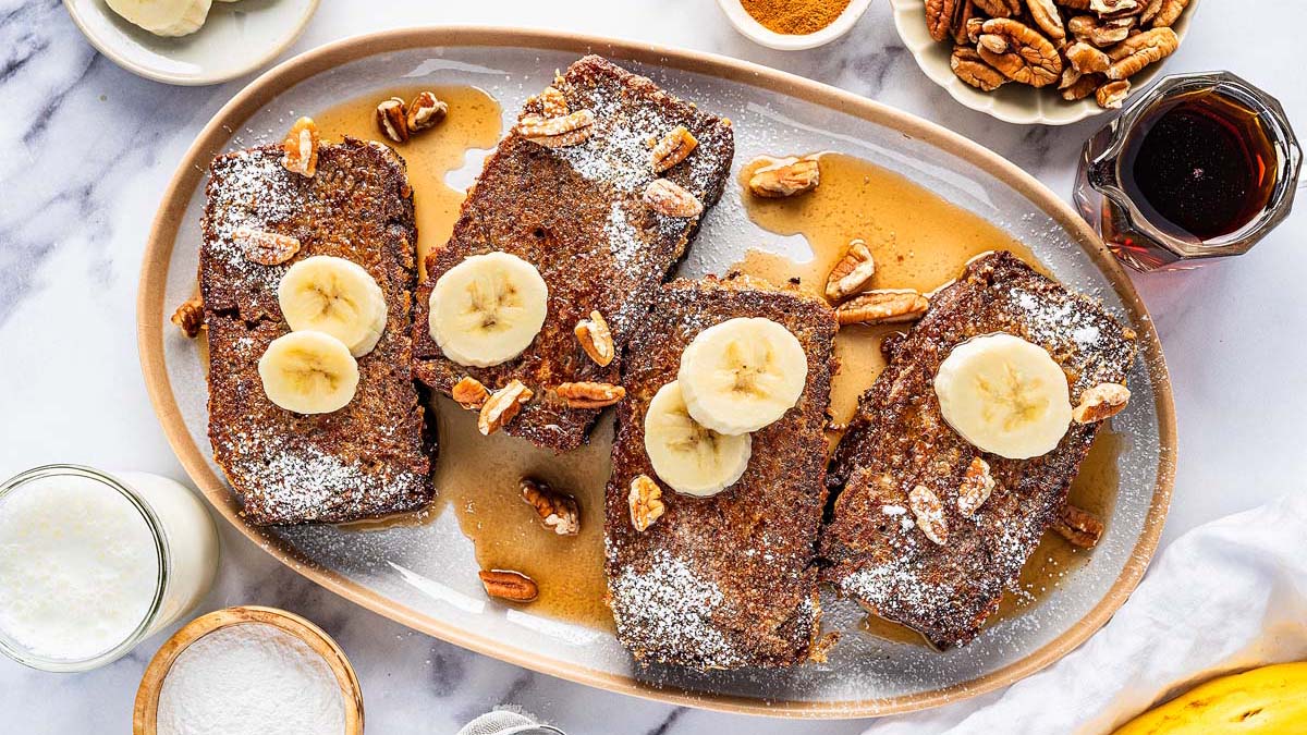 Four slices of French toast topped with banana slices, pecans, powdered sugar, and syrup are served on an oval plate—an easy breakfast perfect for meal prep or trying out new breakfast recipes. Additional toppings and ingredients are nearby.