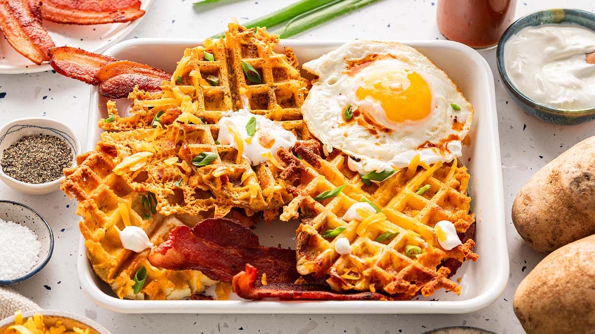 A plate with potato waffles topped with sour cream and green onions, served with crispy bacon and a sunny-side-up egg—perfect for breakfast meal prep. Potatoes and condiments are nearby for easy, healthy breakfast ideas.