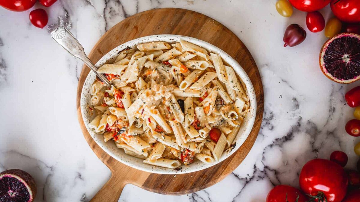 Baked penne pasta with cherry tomatoes and herbs in a white oval dish on a round wooden board, perfect for Sunday dinner and surrounded by tomatoes and a halved blood orange on a marble surface.