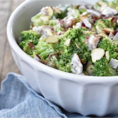 A white bowl filled with broccoli salad, sliced almonds, red onions, and dressing sits on a blue cloth—perfect as one of your favorite side dishes for Sunday dinner.