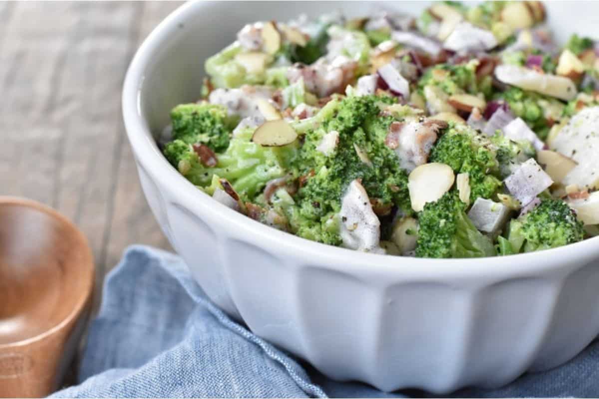 A white bowl filled with broccoli salad, sliced almonds, red onions, and dressing sits on a blue cloth—perfect as one of your favorite side dishes for Sunday dinner.