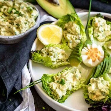 A plate of lettuce boats filled with avocado egg salad, garnished with chives, lemon wedges, and sliced egg—perfect for Easy Dinner Ideas or Kid-Friendly Dinners. A bowl of extra salad and avocado halves are nearby.