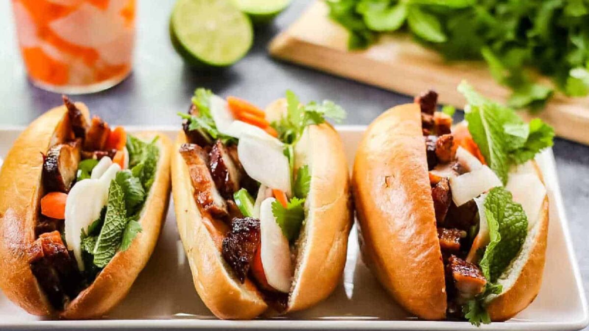 Three bánh mì sandwiches filled with grilled meat, pickled vegetables, fresh herbs, and greens are arranged on a white rectangular plate—perfect inspiration for baked dinner ideas or creative oven recipes.