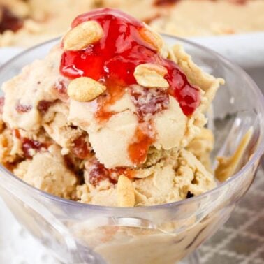 A glass bowl filled with peanut butter ice cream, one of the best frozen treats, is topped with red jelly and a few peanuts, sitting next to a small dish of jelly.