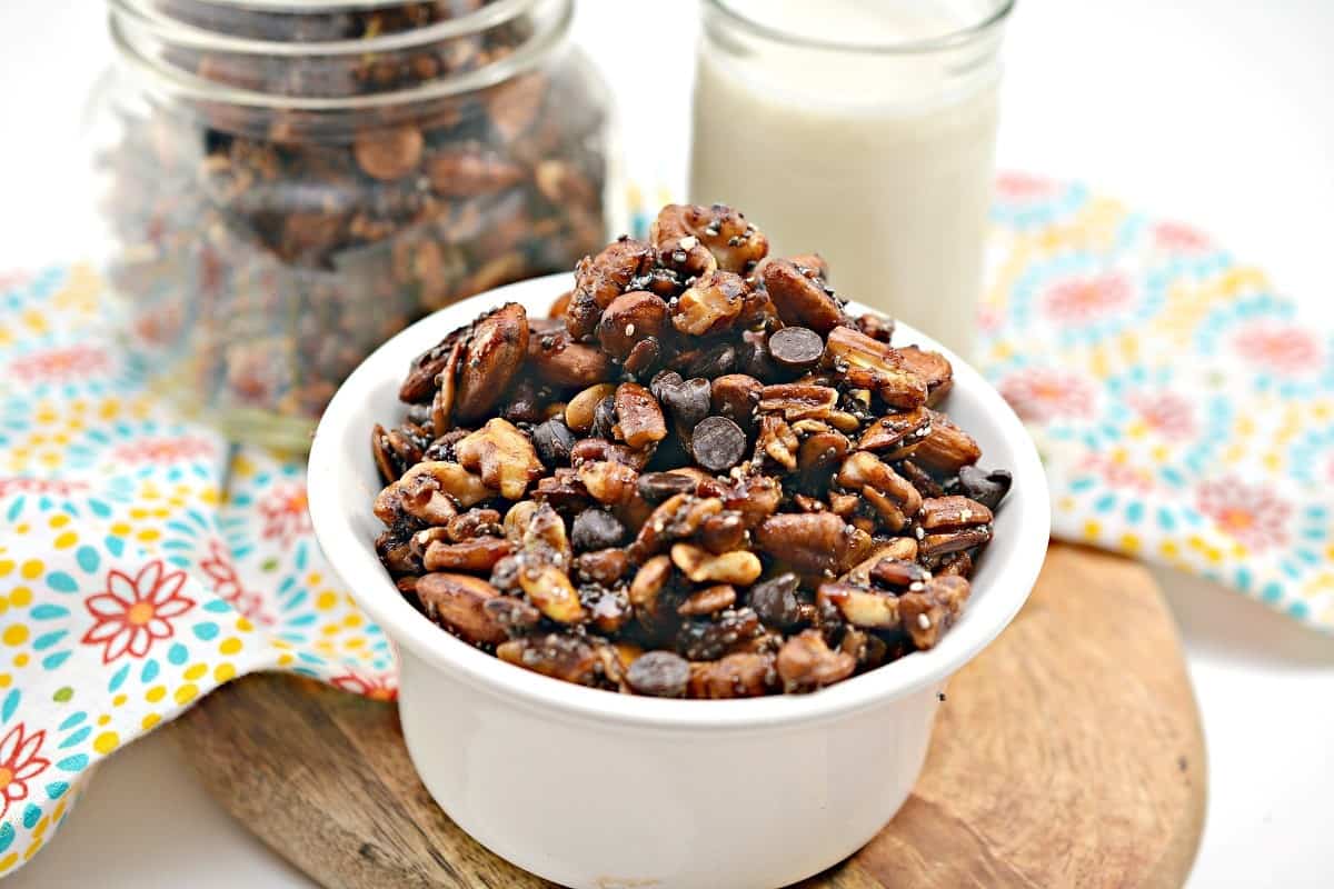 A white bowl filled with mixed nuts and chocolate chips sits on a wooden board, offering unique snacks, with a jar of the same mix and a glass of milk in the background.