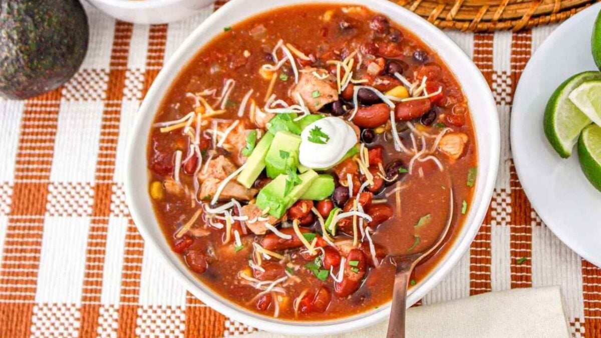A bowl of chili, one of the best easy slow cooker recipes, topped with shredded cheese, avocado, sour cream, and cilantro, with a spoon in the bowl and lime wedges on the side.