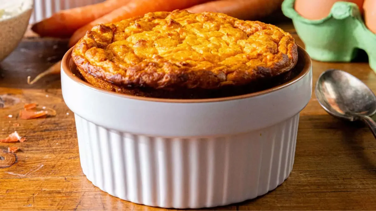 A freshly baked soufflé in a white ramekin sits on a wooden table, surrounded by carrots, eggs, and a spoon—perfect inspiration for new side dishes or recipes at your next main event.