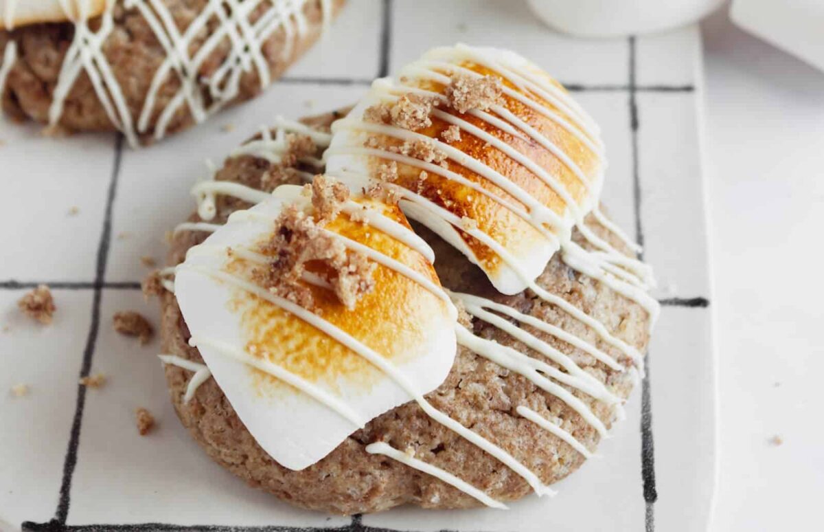 A cookie topped with toasted marshmallows, a sprinkle of crumbs, and drizzled with white icing sits on a white grid-patterned surface—perfect for make-ahead desserts on busy days.
