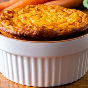 A freshly baked soufflé in a white ramekin sits on a wooden table, surrounded by carrots, eggs, and a spoon—perfect inspiration for new side dishes or recipes at your next main event.