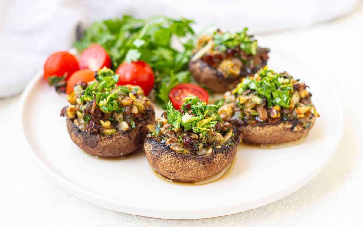 Four stuffed mushrooms topped with chopped herbs are arranged on a white plate, accompanied by cherry tomatoes and fresh parsley—a delicious example of flavorful vegan recipes.