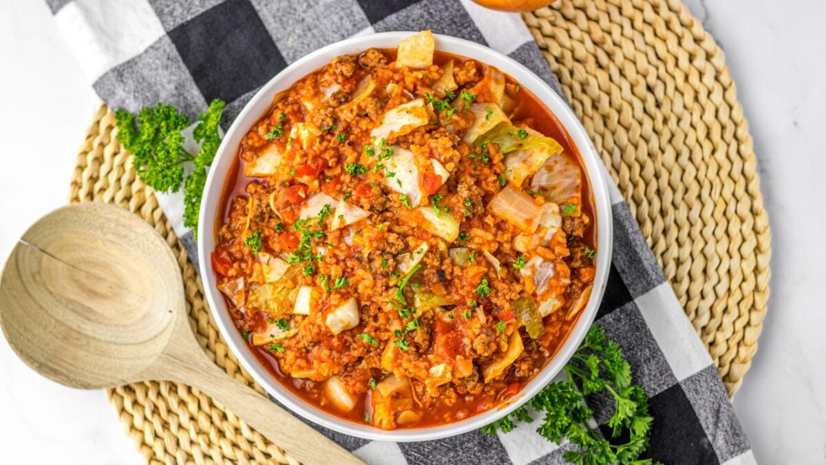 A bowl of cooked cabbage roll casserole with ground meat, rice, and tomato sauce, garnished with parsley, is placed on a woven placemat with a wooden spoon—perfect for fans of Easy Dinner Recipes and Crockpot Meals.