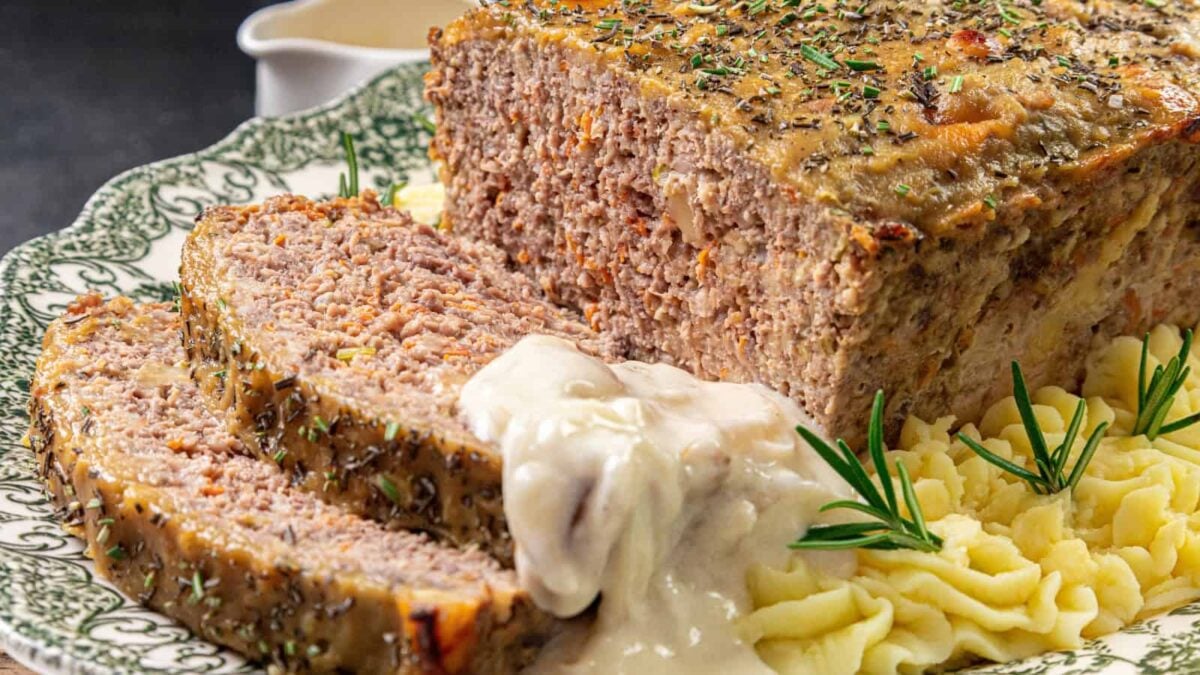 A sliced meatloaf topped with creamy gravy sits on a plate with mashed potatoes, garnished with fresh rosemary and herbs—perfect for easy cooking and simple meals.