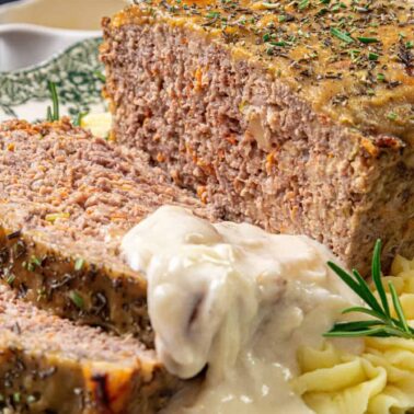 A sliced meatloaf topped with creamy gravy sits on a plate with mashed potatoes, garnished with fresh rosemary and herbs—perfect for easy cooking and simple meals.