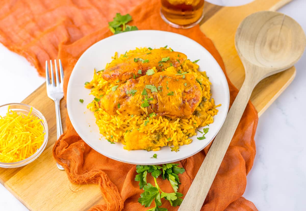 A plate of baked chicken with yellow rice, garnished with chopped parsley, sits on an orange cloth next to a wooden spoon, fork, and a small bowl of shredded cheese—a perfect choice for simple dinner or easy cooking.
