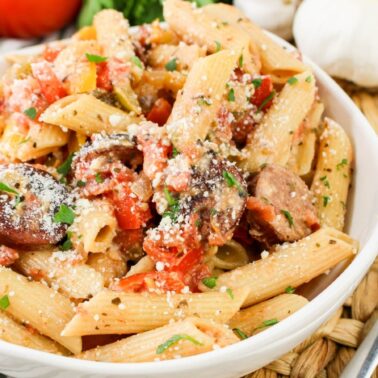 A white bowl filled with penne pasta, sausage slices, diced tomatoes, and sprinkled parmesan cheese sits next to a fork on a woven mat—perfect for anyone seeking delicious and affordable recipes.