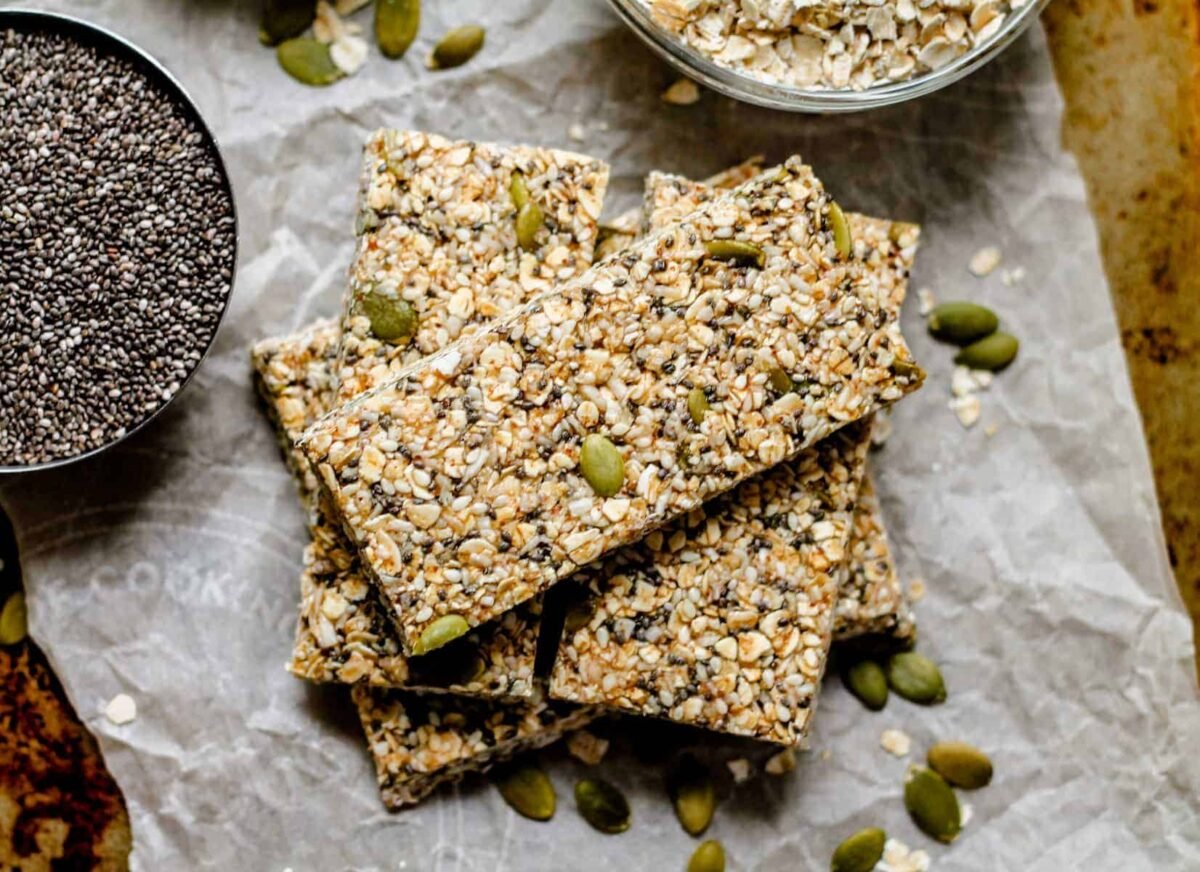A stack of homemade vegan seed and oat bars on parchment paper, with a bowl of oats and a dish of chia seeds nearby. Pumpkin seeds are scattered around, adding extra flavor to these delicious Vegan recipes.