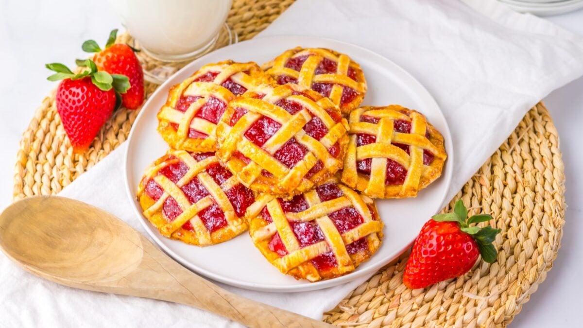 A plate of small lattice-topped strawberry pies is displayed on a woven mat in a cozy kitchen, with fresh strawberries and a glass of milk in the background. A wooden spoon lies nearby.