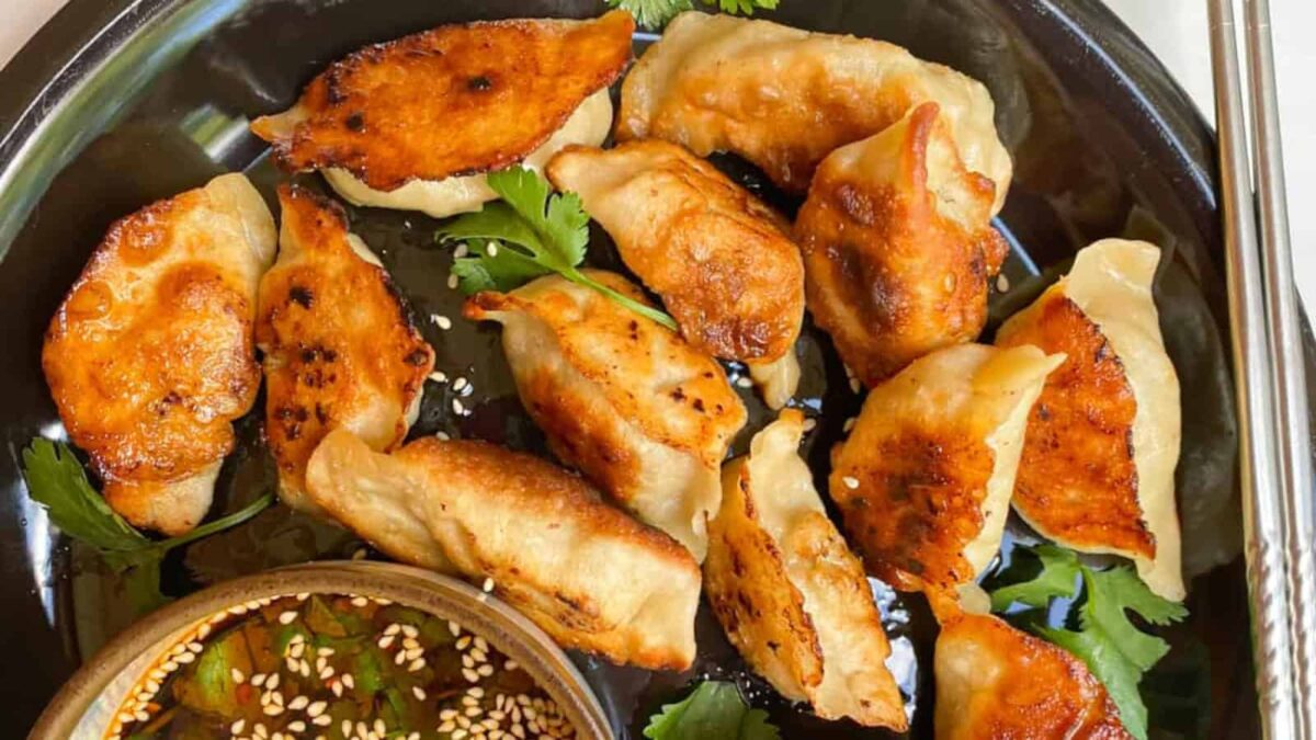 A plate of pan-fried dumplings garnished with cilantro, served with a bowl of soy sauce topped with sesame seeds and vegetables—perfect for those seeking flavorful, dairy-free recipes.