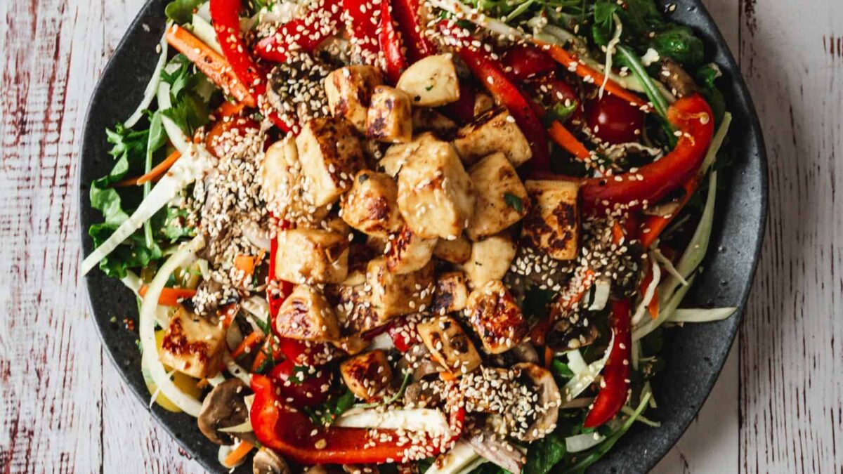 A bowl of salad topped with grilled tofu cubes, sliced red bell peppers, mushrooms, shredded greens, and sprinkled with sesame seeds—perfect for those seeking delicious gluten-free recipes.
