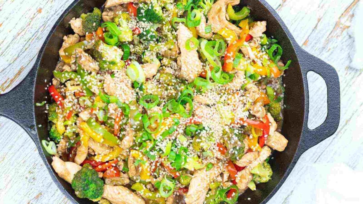 A skillet filled with stir-fried chicken, bell peppers, broccoli, green onions, and sesame seeds on a light wooden surface—a delicious option for those seeking gluten-free recipes.