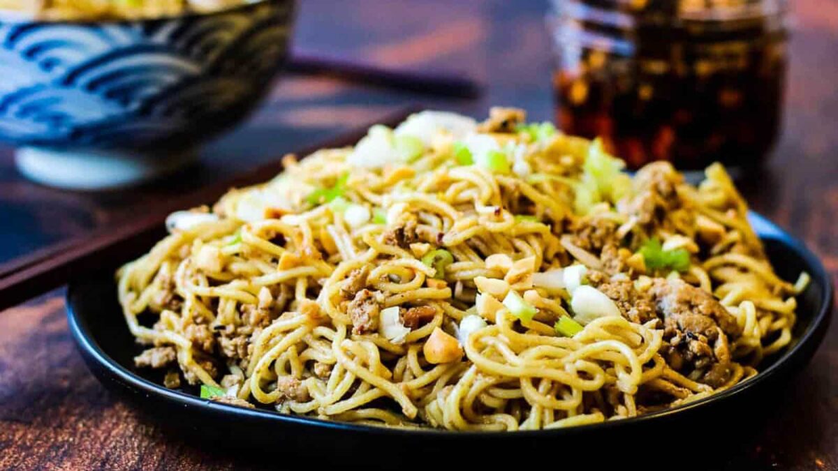A black plate filled with stir-fried noodles, ground meat, chopped nuts, and green onions sits on a wooden table—perfect inspiration for your next gluten-free recipes.