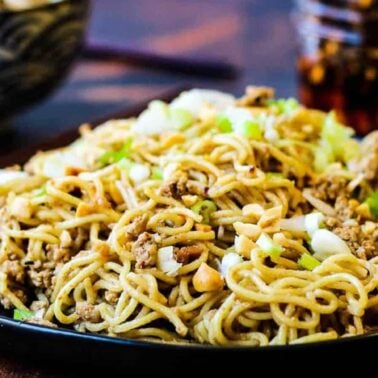 A black plate filled with stir-fried noodles, ground meat, chopped nuts, and green onions sits on a wooden table—perfect inspiration for your next gluten-free recipes.