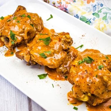 Three pieces of chicken covered in creamy orange sauce, garnished with chopped herbs, are arranged on a white rectangular plate next to a floral-patterned napkin—perfect for easy dinners or quick meals.