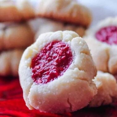 Close-up of shortbread thumbprint cookies with red jam centers, arranged on a red glass plate—perfect for anyone seeking flavorful vegan recipes. More cookies are stacked in the background, making this treat ideal for plant-based dessert lovers.