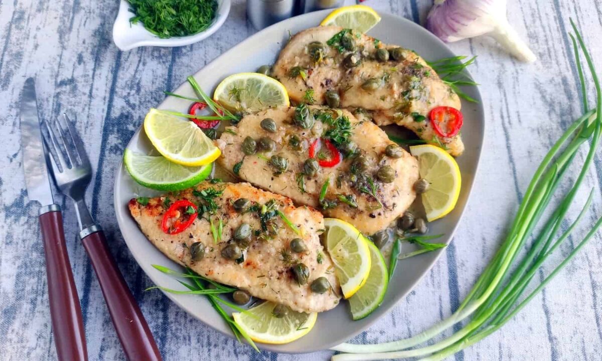 Three cooked chicken breasts garnished with capers, lemon slices, red chili, and fresh herbs on a plate—perfect for easy chicken recipes or weeknight dinners. Green onions and cutlery are placed nearby on a light wooden surface.