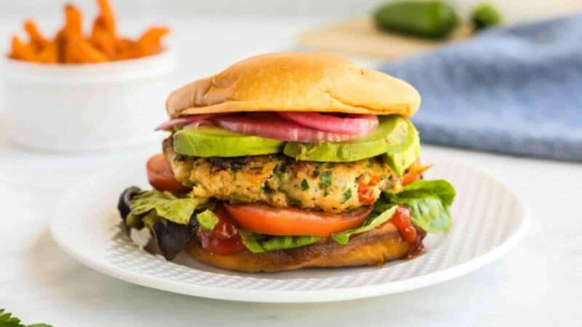 A veggie burger on a white plate, topped with sliced avocado, tomato, lettuce, and pickled onions—bringing a fresh twist to Thanksgiving classics—with sweet potato fries on the side.