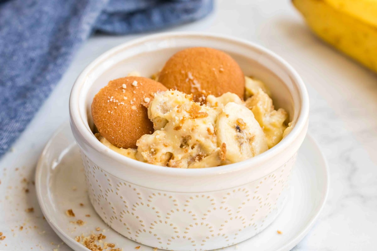 A small white ramekin filled with delicious banana pudding, topped with banana slices and two vanilla wafer cookies, sits on a white plate—a perfect addition to your collection of banana treats and recipes.