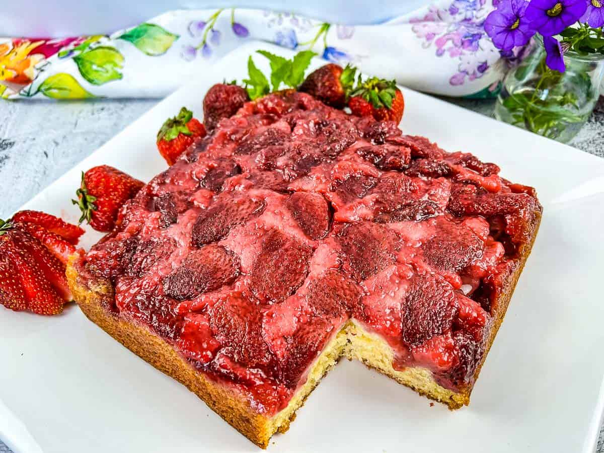 A square strawberry upside-down cake with a slice missing, garnished with fresh strawberries, on a white plate next to a floral napkin and purple flowers—perfect for busy days or as one of your favorite make-ahead desserts.
