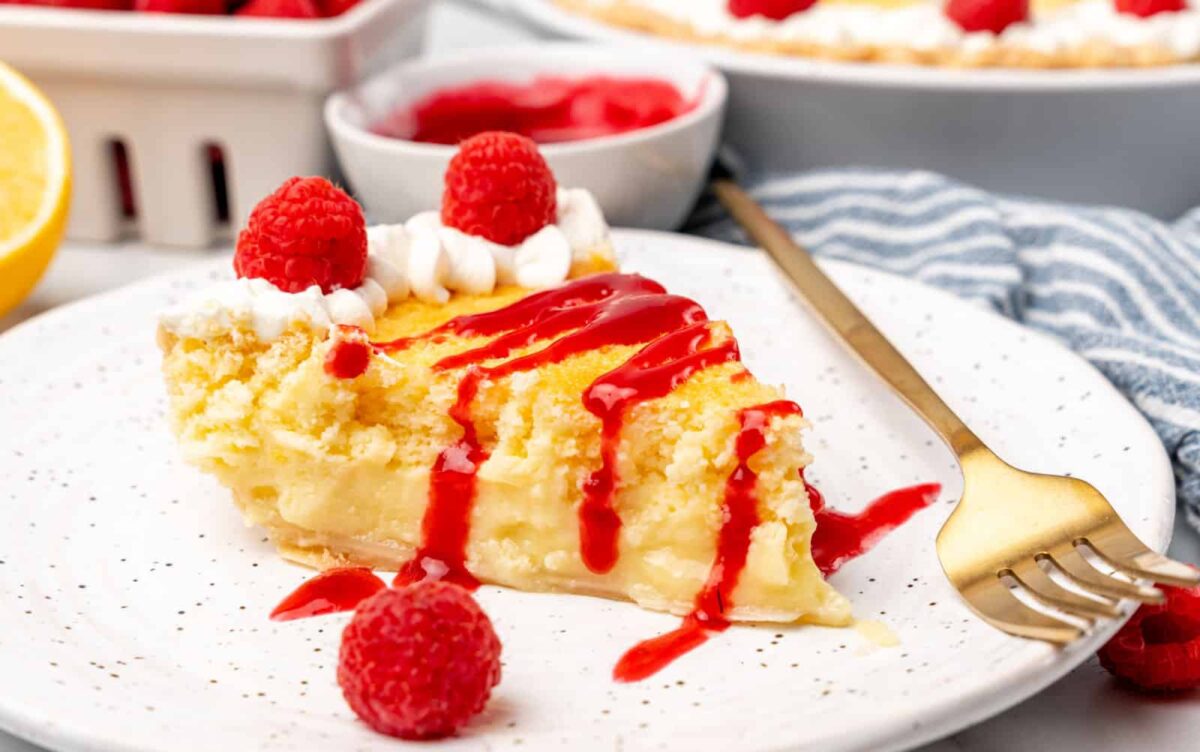 A slice of creamy pie inspired by Lemon Desserts, topped with whipped cream, raspberries, and raspberry sauce sits on a white plate with a gold fork.