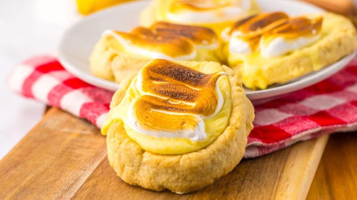 A close-up of lemon meringue cookies, a no bake treat among easy recipes for lemon desserts, with browned meringue topping, arranged on a wooden board and a white plate over a red checkered cloth.